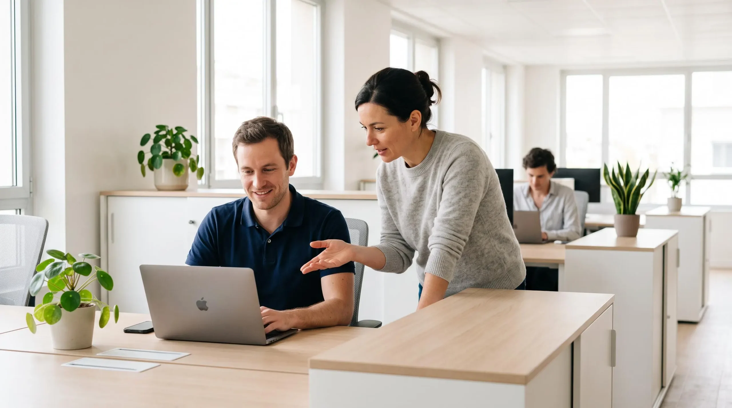 Nouveau collaborateur à son poste de travail accompagné par une collègue expérimentée lui montrant des informations sur l'écran dans un open space lumineux et accueillant